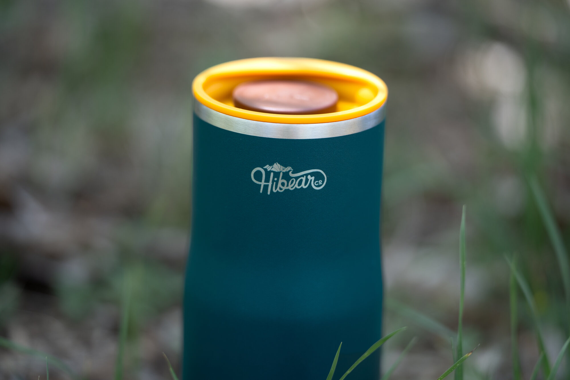 Close-up of a teal Hibear adventure flask with its yellow rim and logo, grass behind