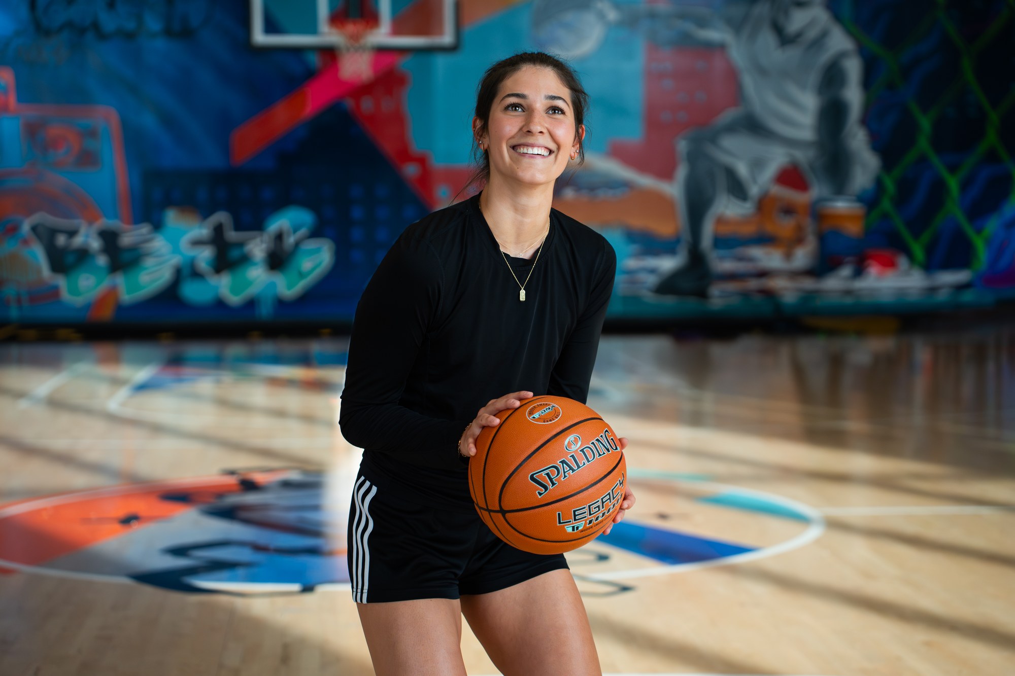 Smiling player holding a Spalding Legacy basketball on an indoor court with a mural wall