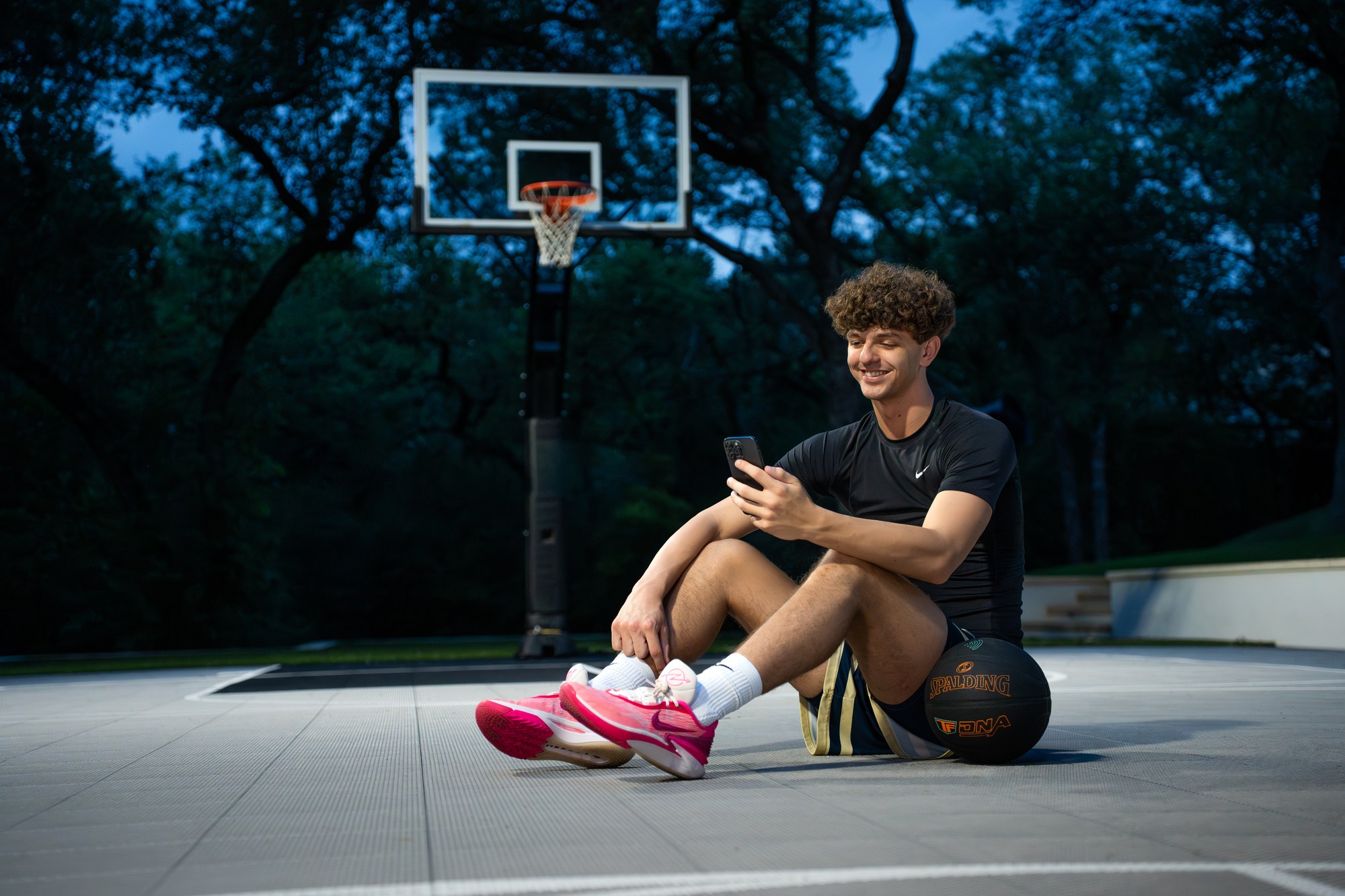 Player sitting on an outdoor court at dusk checking his phone, black Spalding TF DNA ball beside him